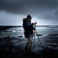 Man in Coat and Hat setting up Tripod on Beach under clouds